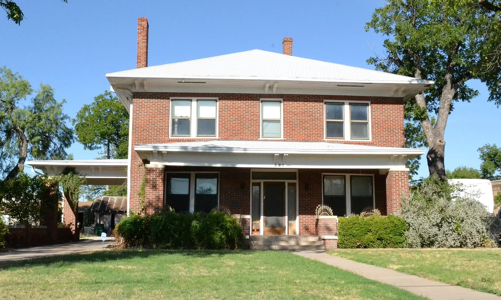 Red brick two-story Texas home with a hipped roof, front porch and live oaks on a sunny day