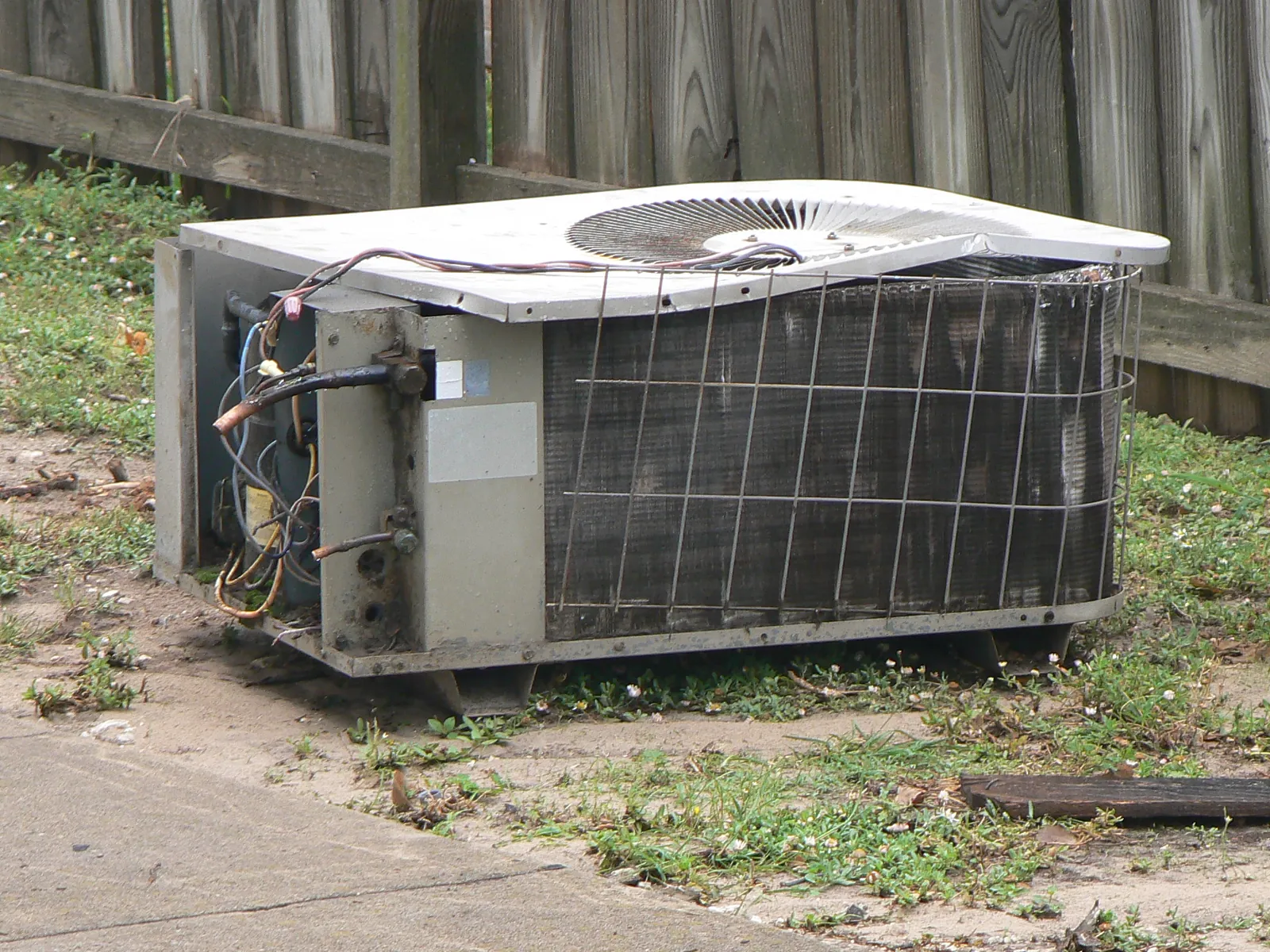 Outdoor AC condenser with its service panel off and the wiring exposed for repair, beside a wood privacy fence